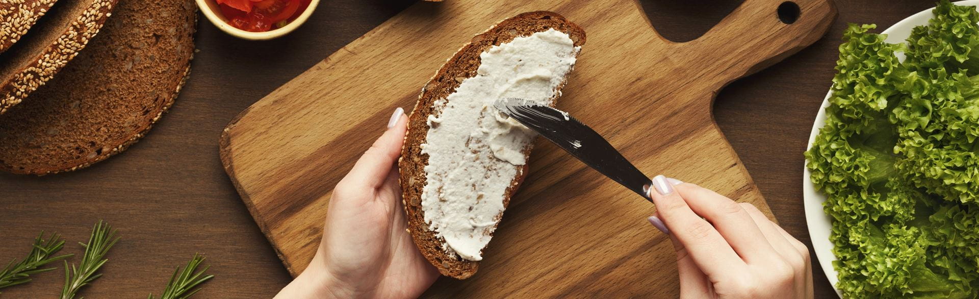 Person spreading a creamy white spread on brown bread, surrounded by fresh vegetables and herbs on a wooden board.