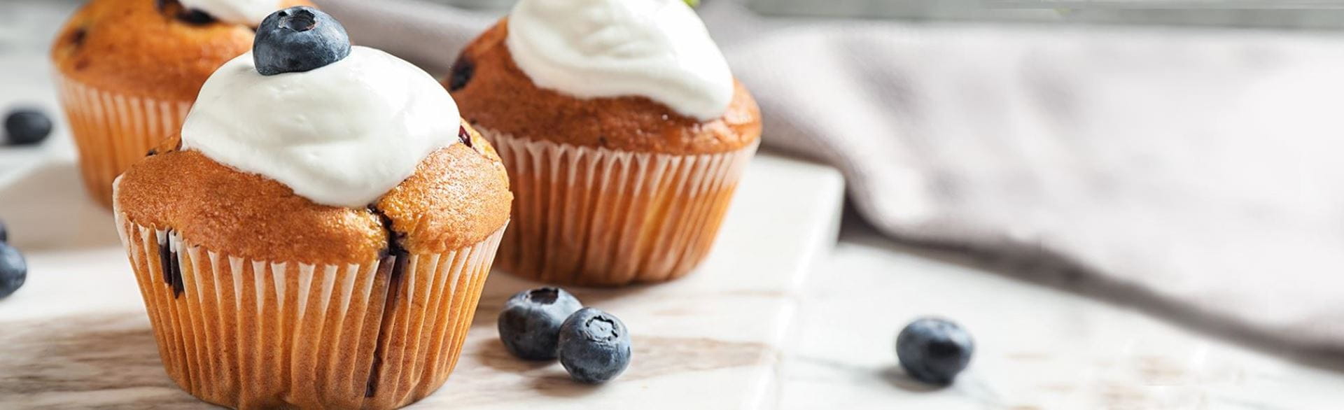 Three blueberry muffins with whipped cream and blueberries on a white surface
