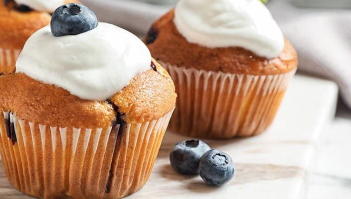 Three blueberry muffins with cream and blueberries, surrounded by loose berries